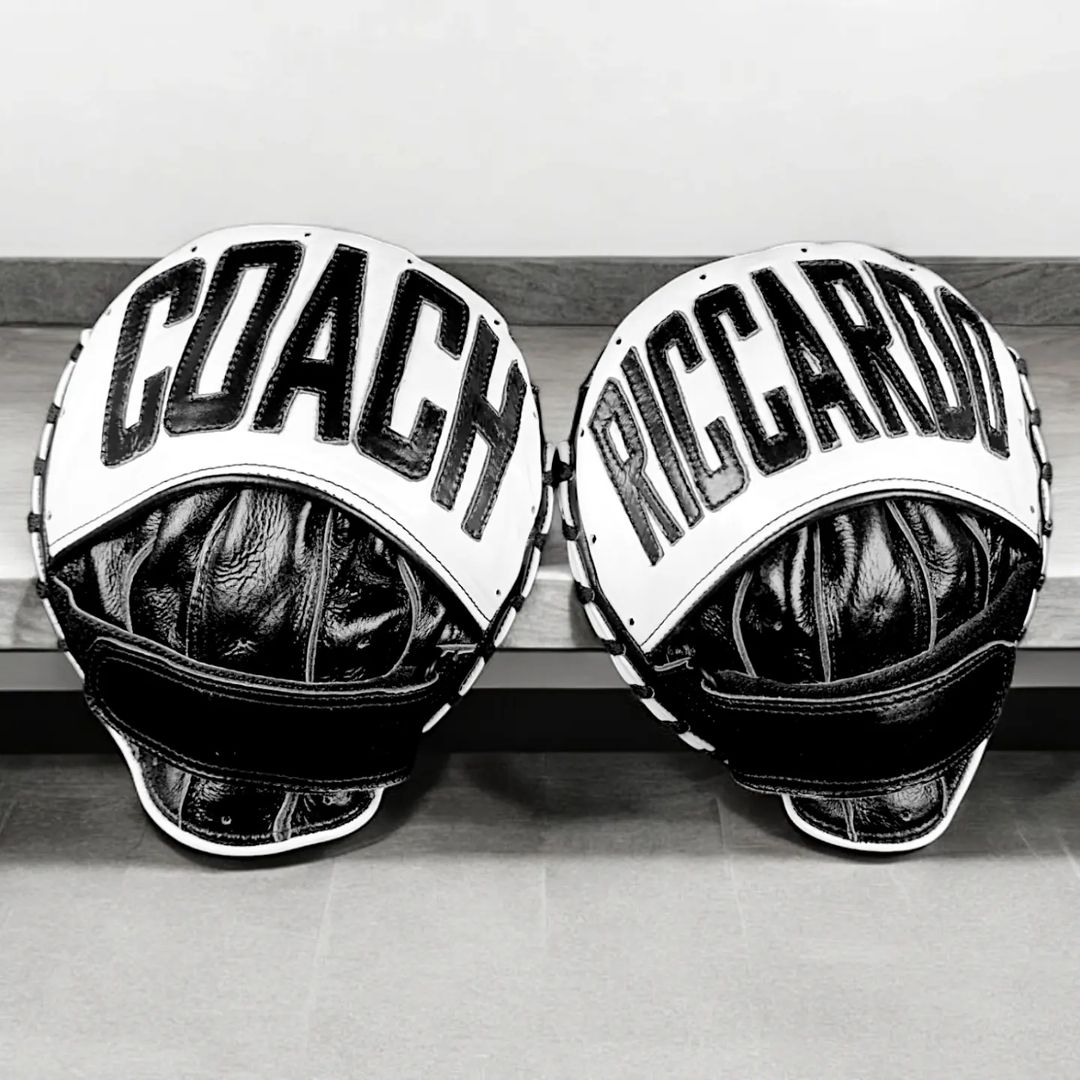 Two black and white boxing pads rest on a bench. One pad is labeled COACH and the other RICCARDO in bold letters.