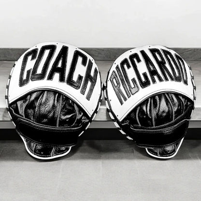 Two black and white personalised boxing gloves, each custom embroidered—one with "COACH," the other with "RICCARDO" in bold uppercase—sit side by side on a bench. Perfect personalized gifts for boxers and sports lovers.